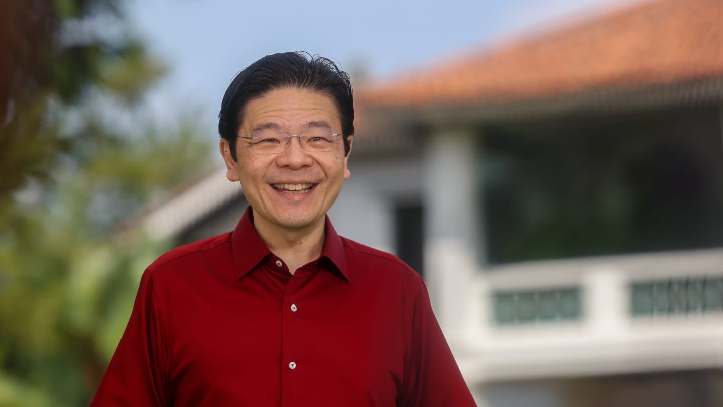 Man in red shirt and glasses smiling, standing outside a white house with a red tiled roof.